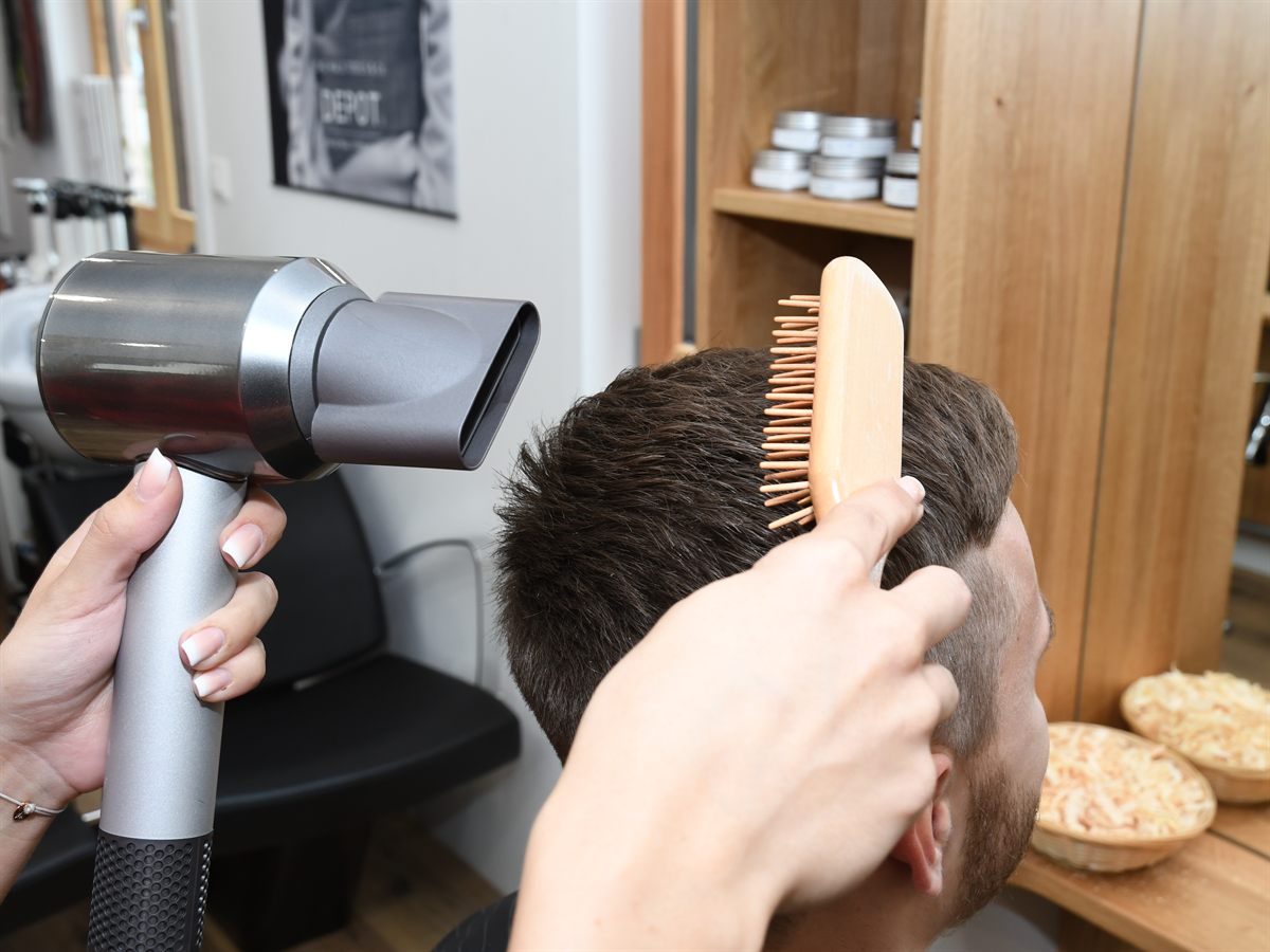 Nahaufnahme: eine Hand hält den Föhn, die andere eine Holzbürste im kurzen Haar eines Kunden.