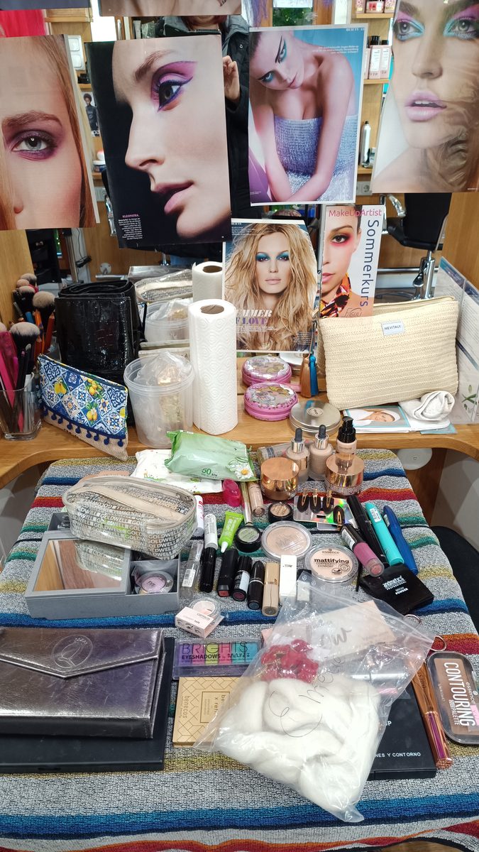 Ein Tisch voller Schminkpaletten, Pinsel, Puder und Haarnadeln, dahinter Poster mit Make-up-Motiven.