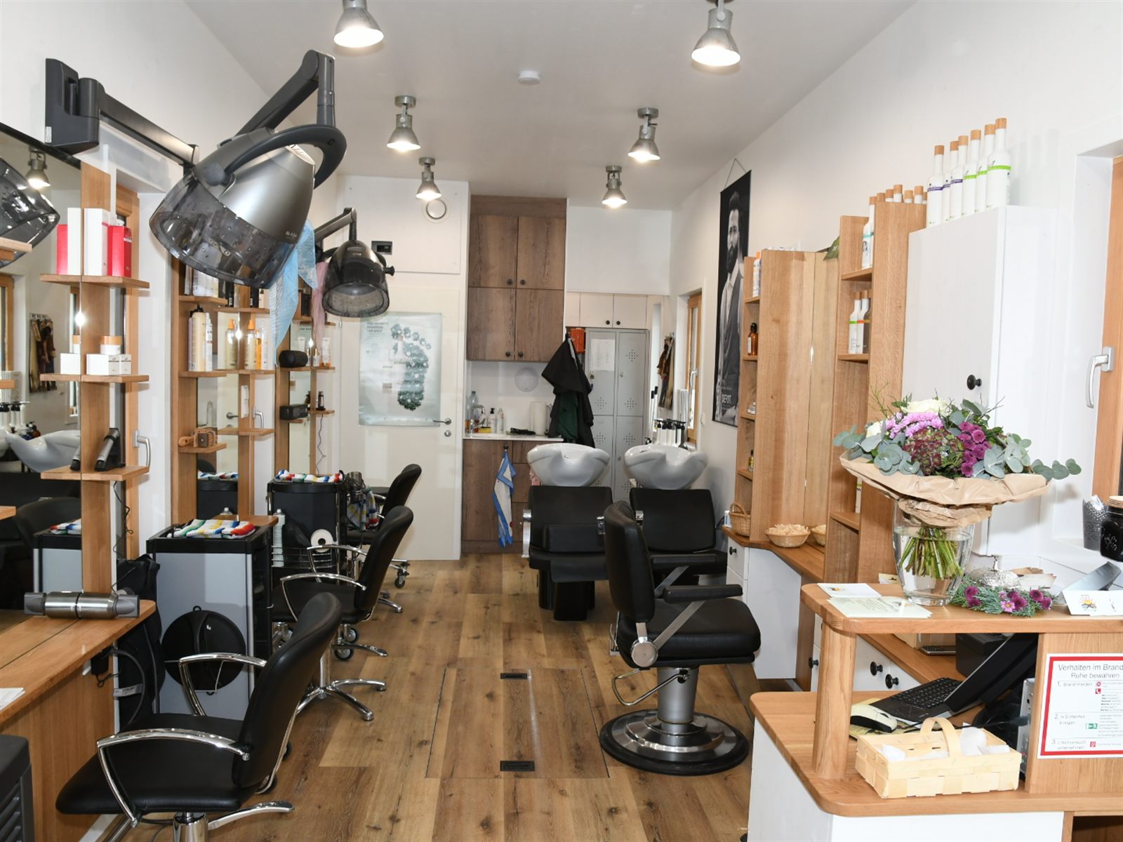 Blick durch den Salon: helle Eichenmöbel, Spiegel, zwei Waschplätze, Friseurstühle und Produktregale, hinten eine Tür ins Nebenzimmer.