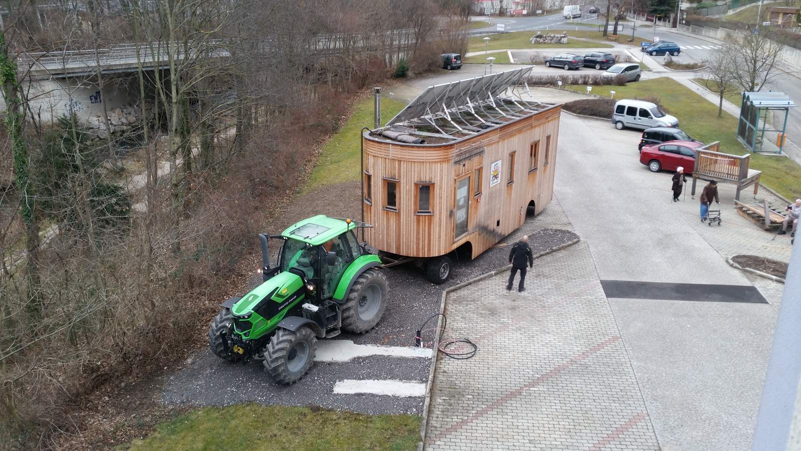 Der Traktor zieht den Wagon über einen gepflasterten Platz, im Hintergrund geparkte Autos und bewaldete Hänge.