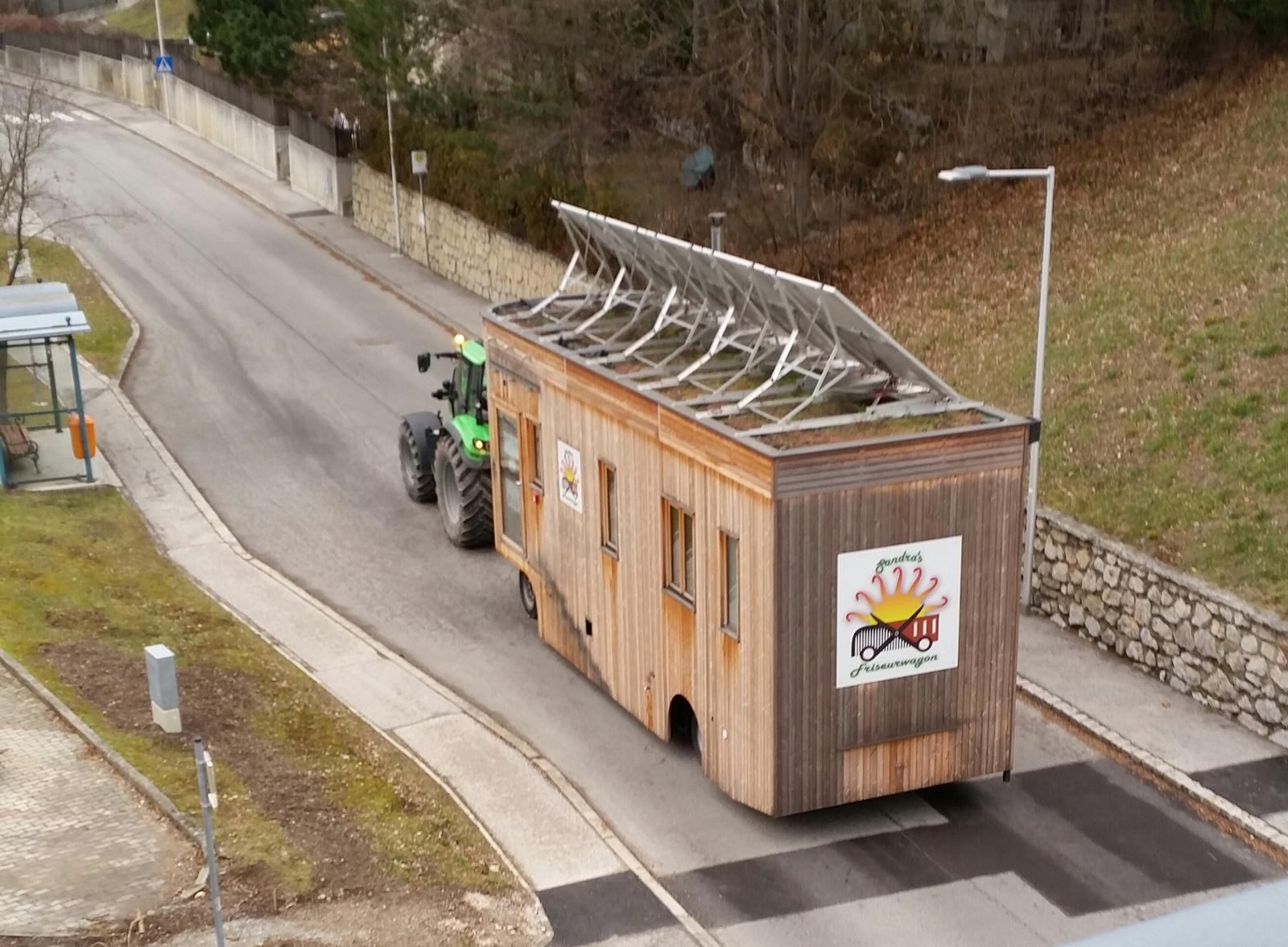 Der Wagon hängt an einem grünen Traktor und fährt eine Ortsstraße entlang, auf dem Dach das leere Gestell der Solarmodule.