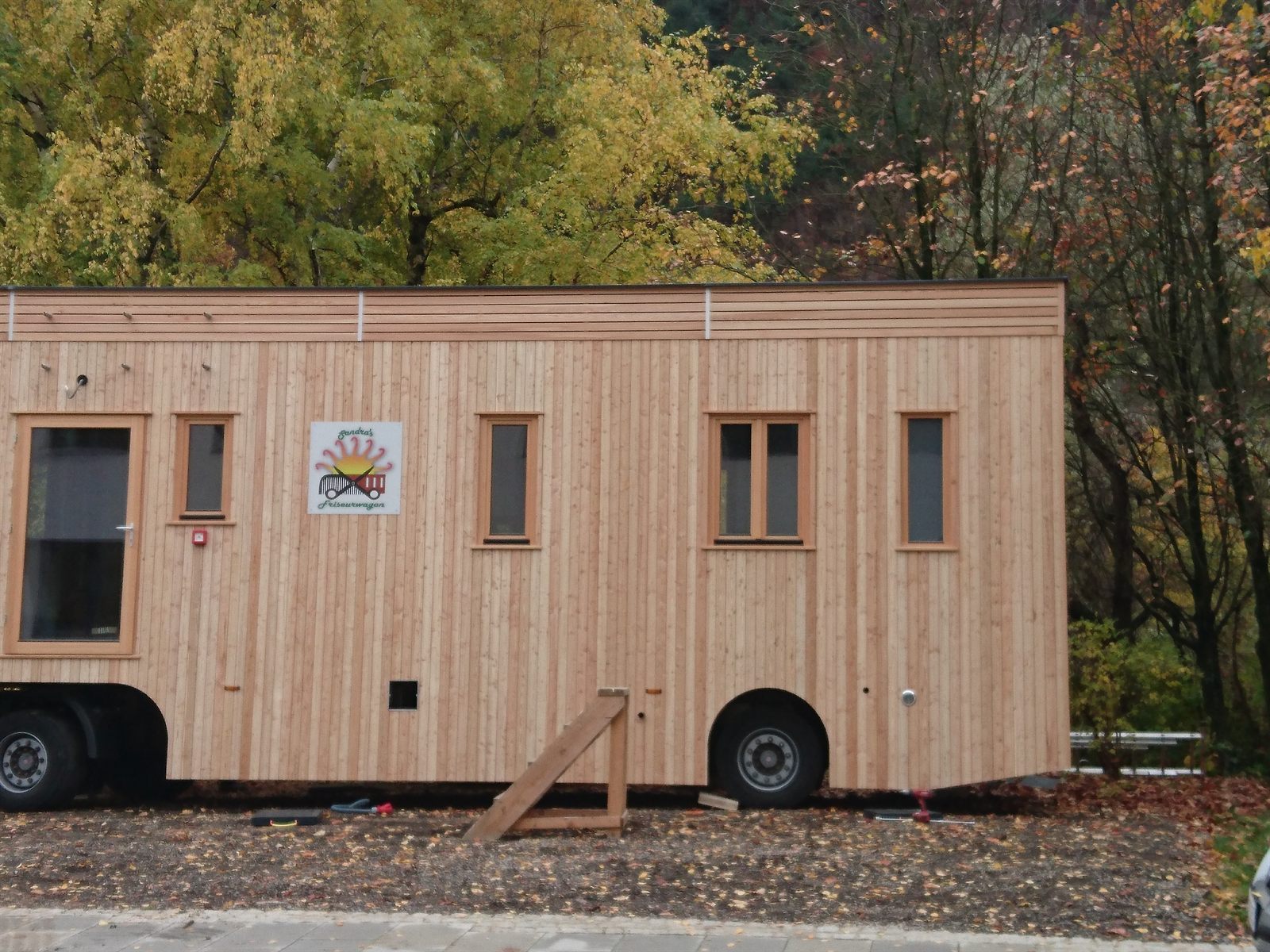 Der Friseurwagon von der Seite: senkrechte Lärchenholzschalung, sechs Fenster, das Logo an der Wand, dahinter herbstlicher Laubwald.