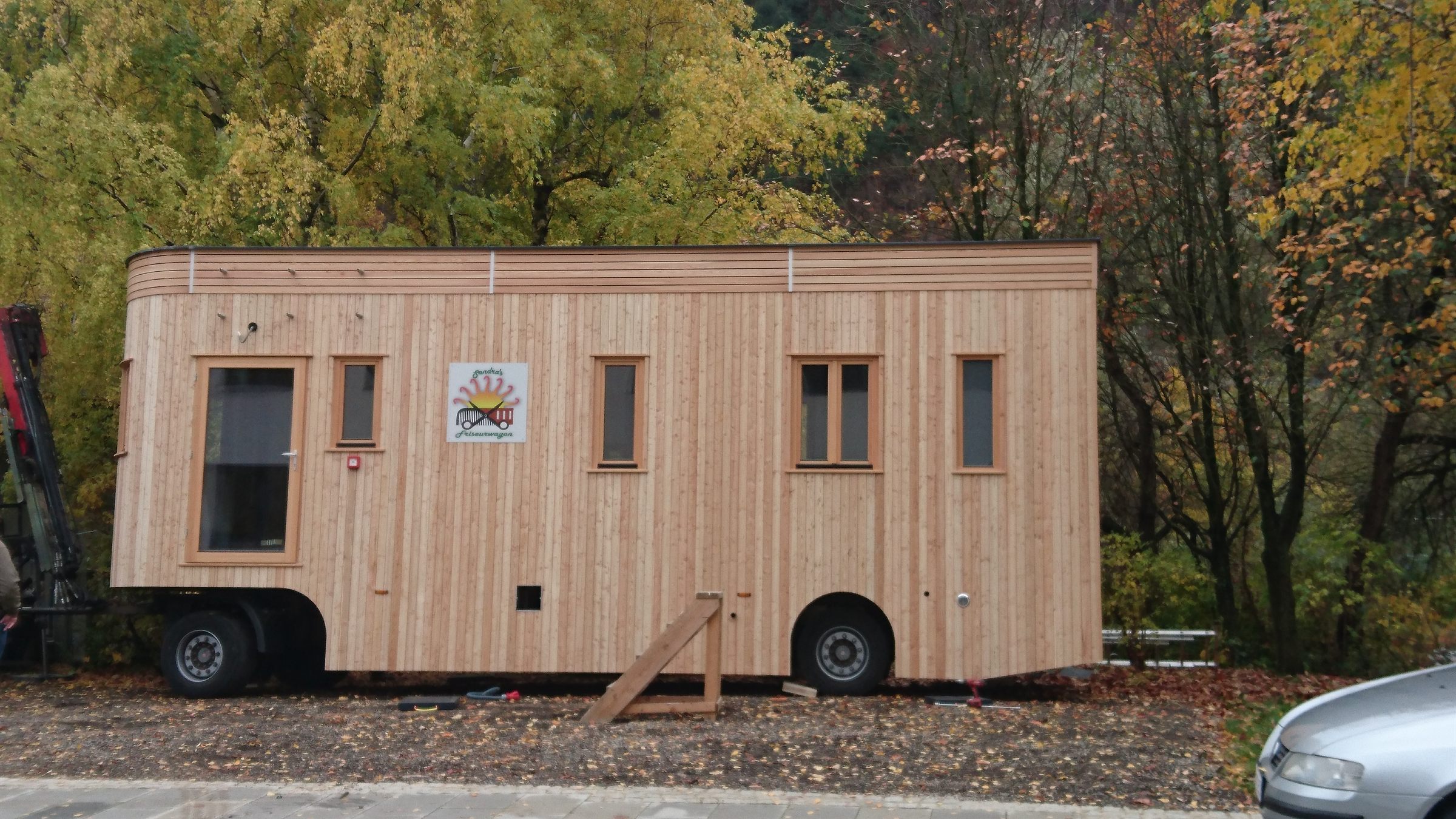 Der Friseurwagon von der Seite: ein langer Wagon mit senkrechter Lärchenholzschalung, sechs Fenstern und dem Logo an der Wand, dahinter herbstlicher Laubwald.